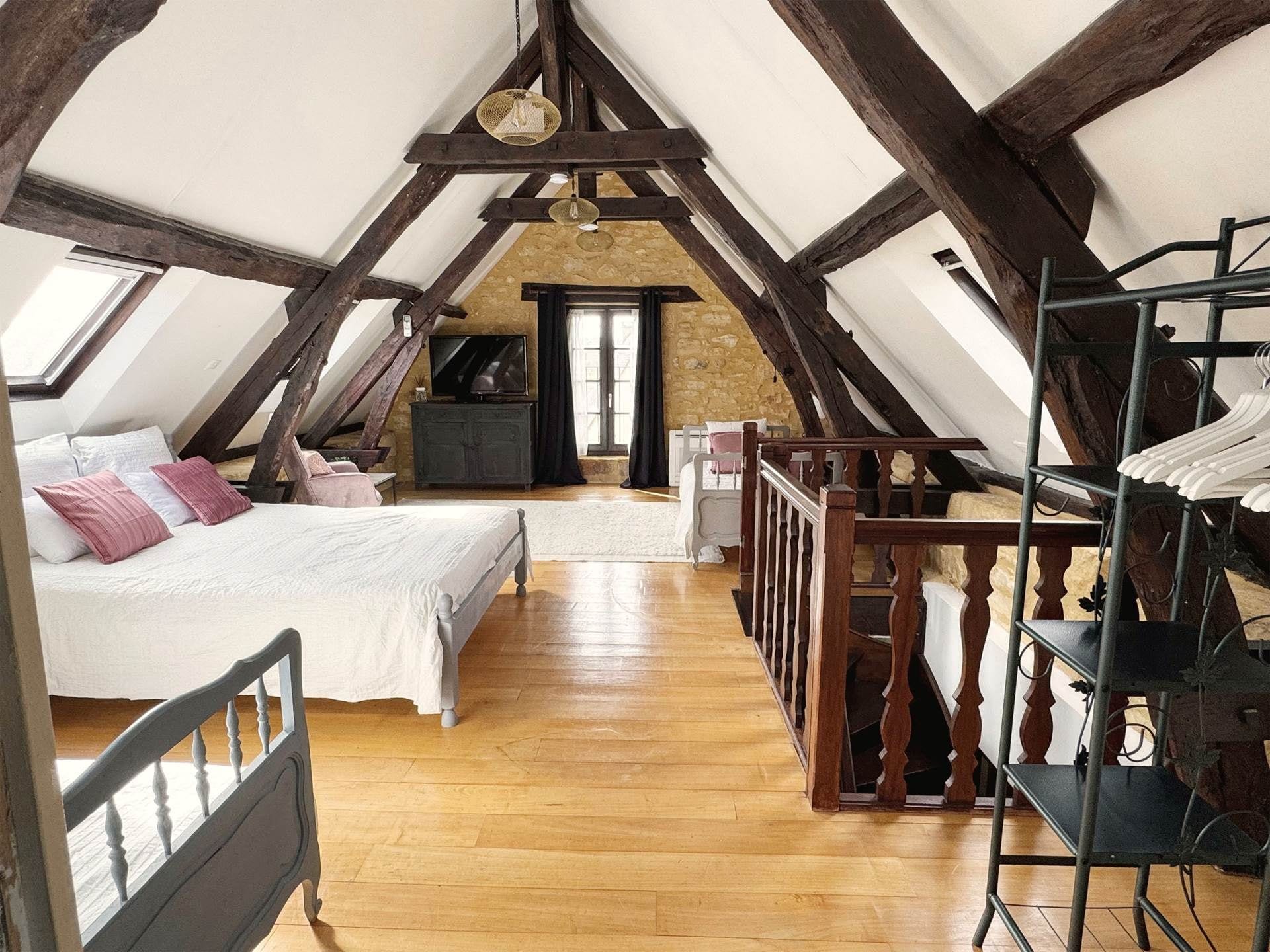 Un dormitori tipus loft amb sostre inclinat, bigues de fusta, un llit, una petita arca, un televisor i una estant per a la roba.