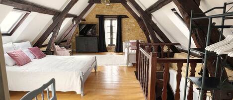 A loft bedroom with a sloped ceiling, wooden beams, a bed, a dresser, a TV, and a clothing rack.