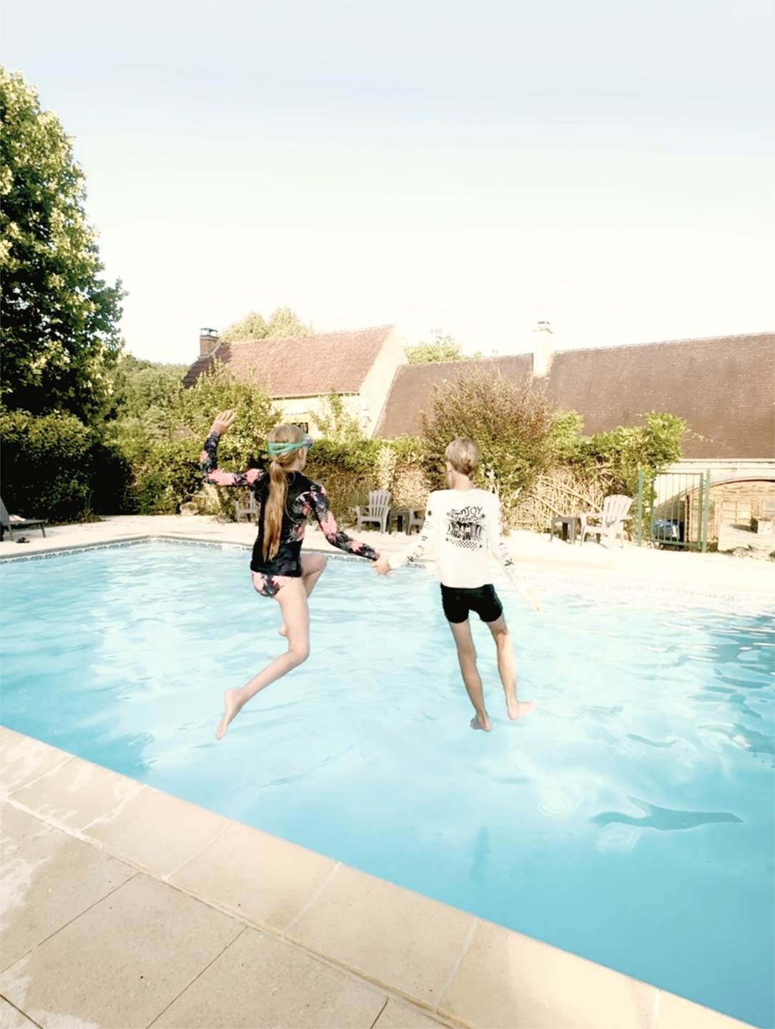 Ein Pool mit einer gefliesten Kante, umgeben von Grünflächen und einem Gebäude mit einem geneigten Dach.
