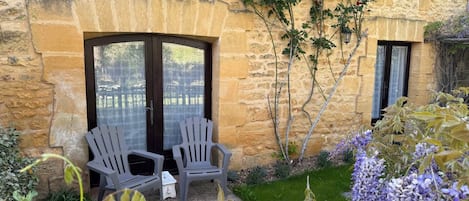 A stone building with two dark-framed glass doors, a small garden with purple flowers, and two grey chairs.