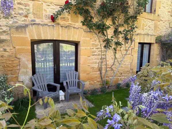 Un edificio de piedra con dos puertas de vidrio con marco oscuro, un pequeño jardín con flores moradas y dos sillas grises.
