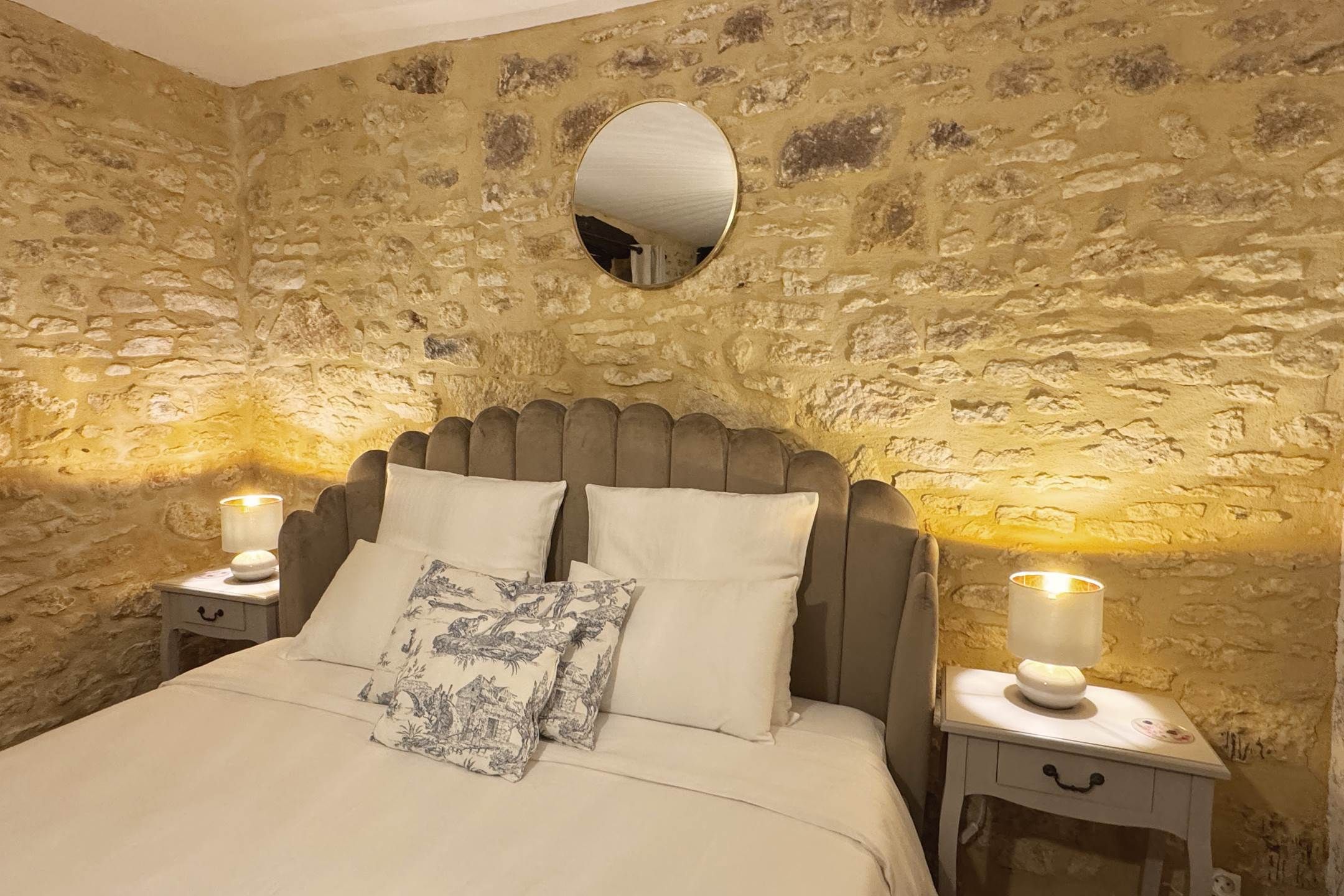 A bedroom with a stone wall, a round mirror, a bed with two pillows, and bedside lamps.