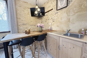 A kitchen with a stone wall, wooden bar stools, a wooden table, a hanging light fixture, a vase with flowers, and a sink. - La Campanule – Spacious holiday home for 6 people with private hot tub (Meyrals)