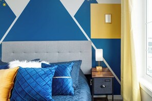 A bedroom with a bed, pillows, a nightstand, a lamp, and a wall with geometric patterns. - ~PrivateFencedYard | 2FamilyRms | SunshineYellow (Calgary)