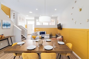 A modern dining area with a wooden table set for four, a sofa in the background, and a staircase leading to an upper level. - ~PrivateFencedYard | 2FamilyRms | SunshineYellow (Calgary)