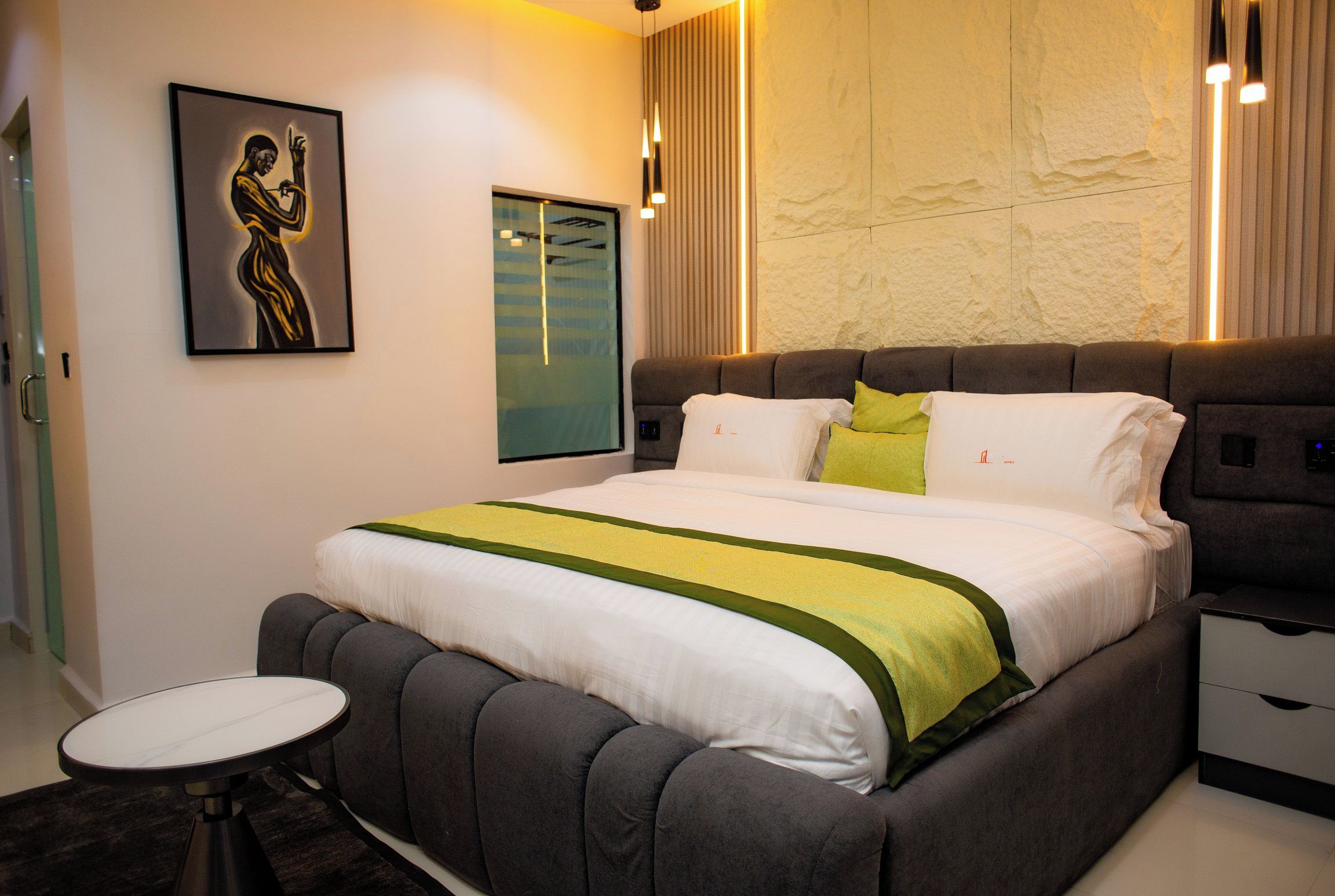 A modern bedroom with a large bed, bedside tables, a round side table, and a wall-mounted artwork.