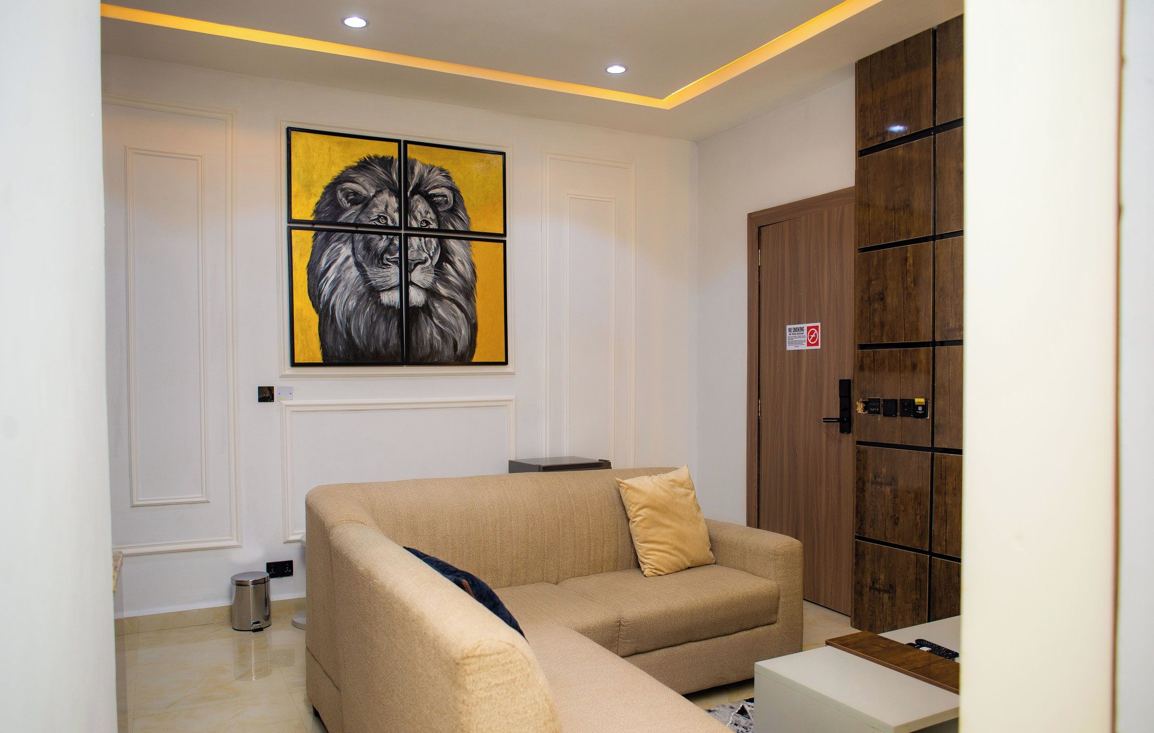 A living room with a beige sofa, a painting of a lion's face, a wooden wardrobe, and a 'No Smoking' sign.