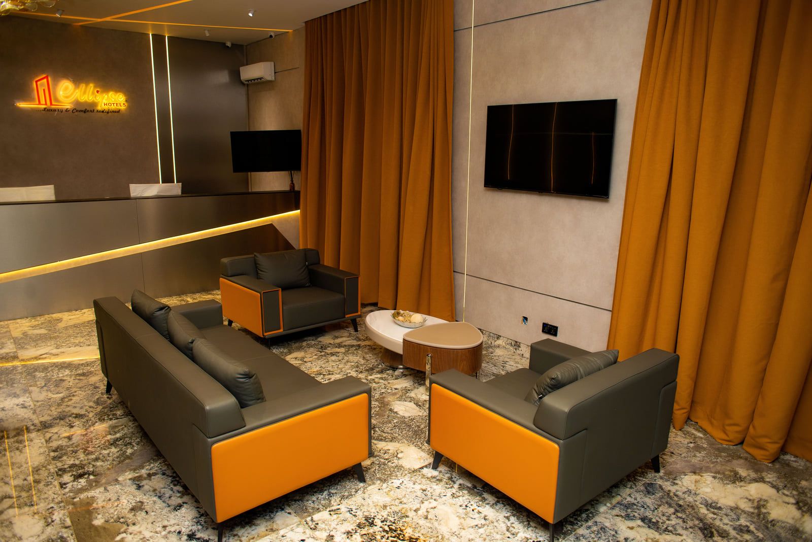 A modern hotel lobby with a seating area featuring sofas and armchairs, a flat-screen TV mounted on the wall, and a decorative wall with illuminated signage.