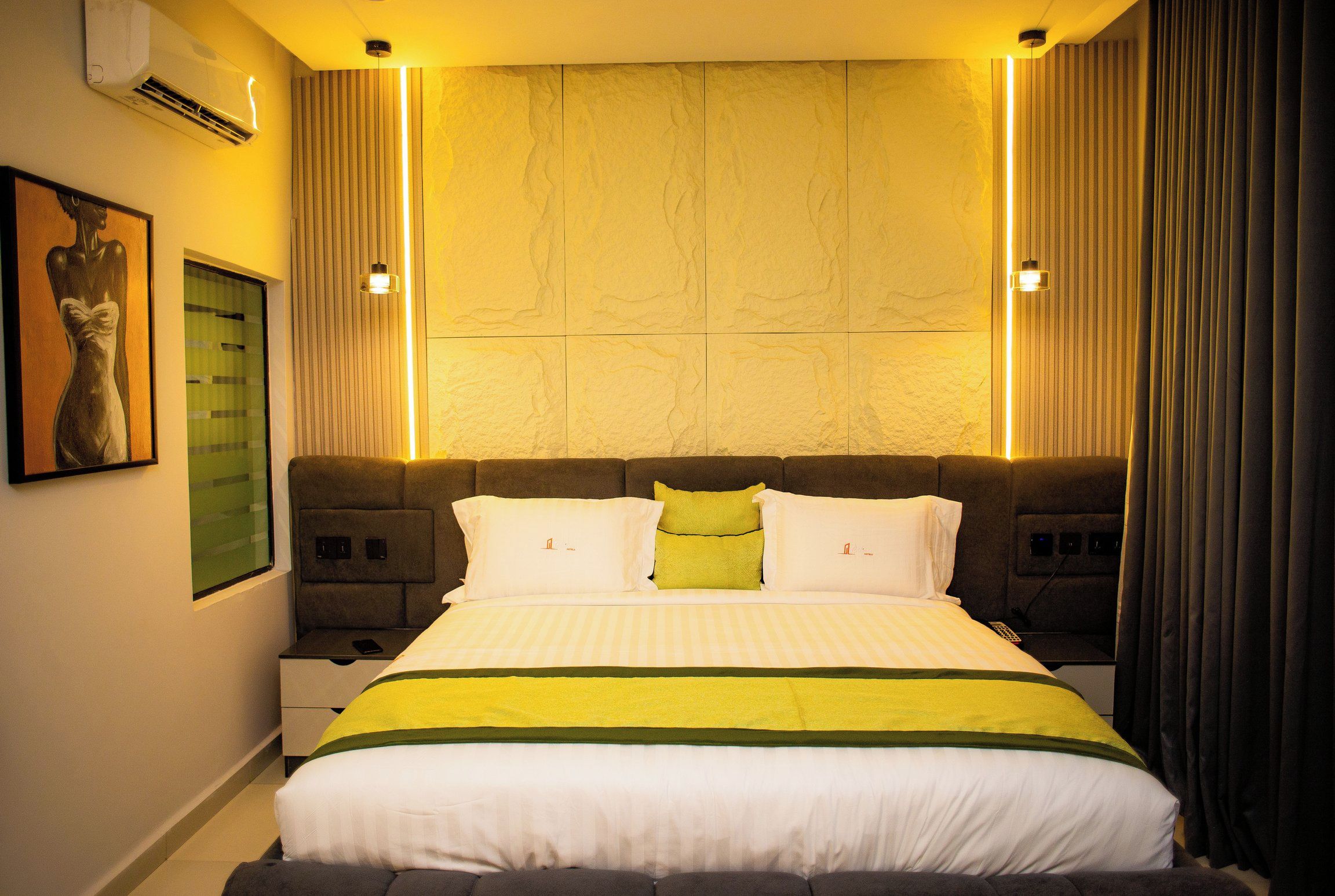 A hotel room with a large bed, a headboard with built-in lighting, a wall-mounted air conditioner, and a framed artwork on the wall.