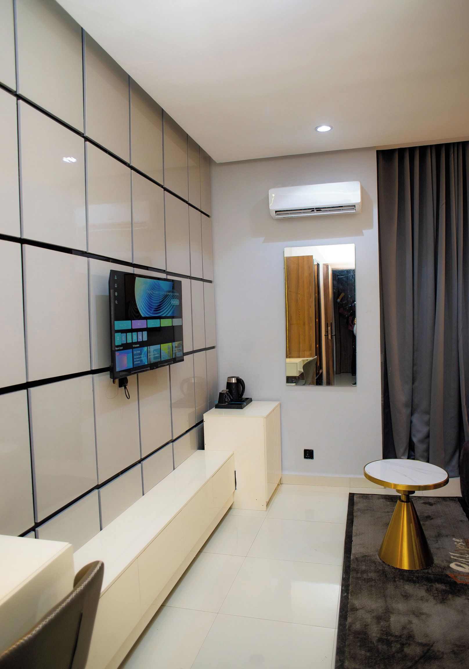 Una sala de estar moderna con un televisor de pantalla plana montado en una pared de azulejos, una cafetera sobre una consola blanca y una mesa redonda con base dorada.