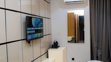 Una sala de estar moderna con un televisor de pantalla plana montado en una pared de azulejos, una cafetera sobre una consola blanca y una mesa redonda con base dorada.