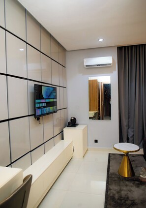 A modern living room with a flat-screen TV mounted on a tiled wall, a coffee machine on a white console table, and a round table with a gold base. - Ellipse Hotels (Lagos)