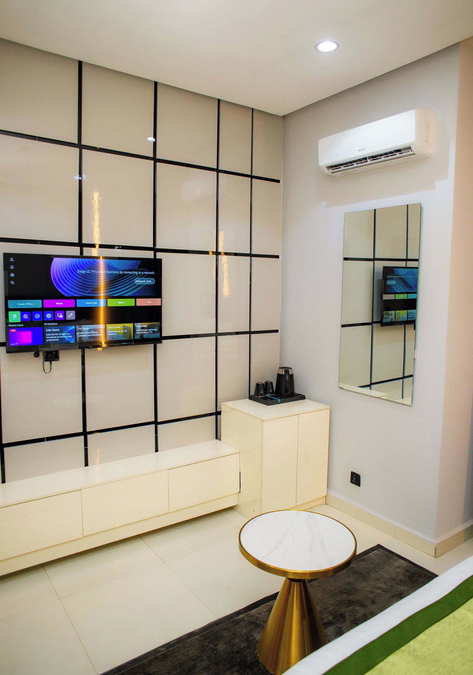 A modern living room with a flat-screen TV mounted on a wall with a grid pattern, a coffee machine on a white cabinet, a round table with a marble top, and a large mirror on the wall.