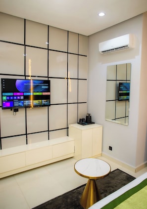A modern living room with a flat-screen TV mounted on a wall with a grid pattern, a coffee machine on a white cabinet, a round table with a marble top, and a large mirror on the wall. - Ellipse Hotels (Lagos)