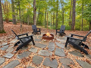 A fire pit surrounded by four Adirondack chairs on a stone-paved area with a forest backdrop. - Doe Run Hideaway - Pet Friendly | Hot Tub | Game Room | Firepit (Ellijay)