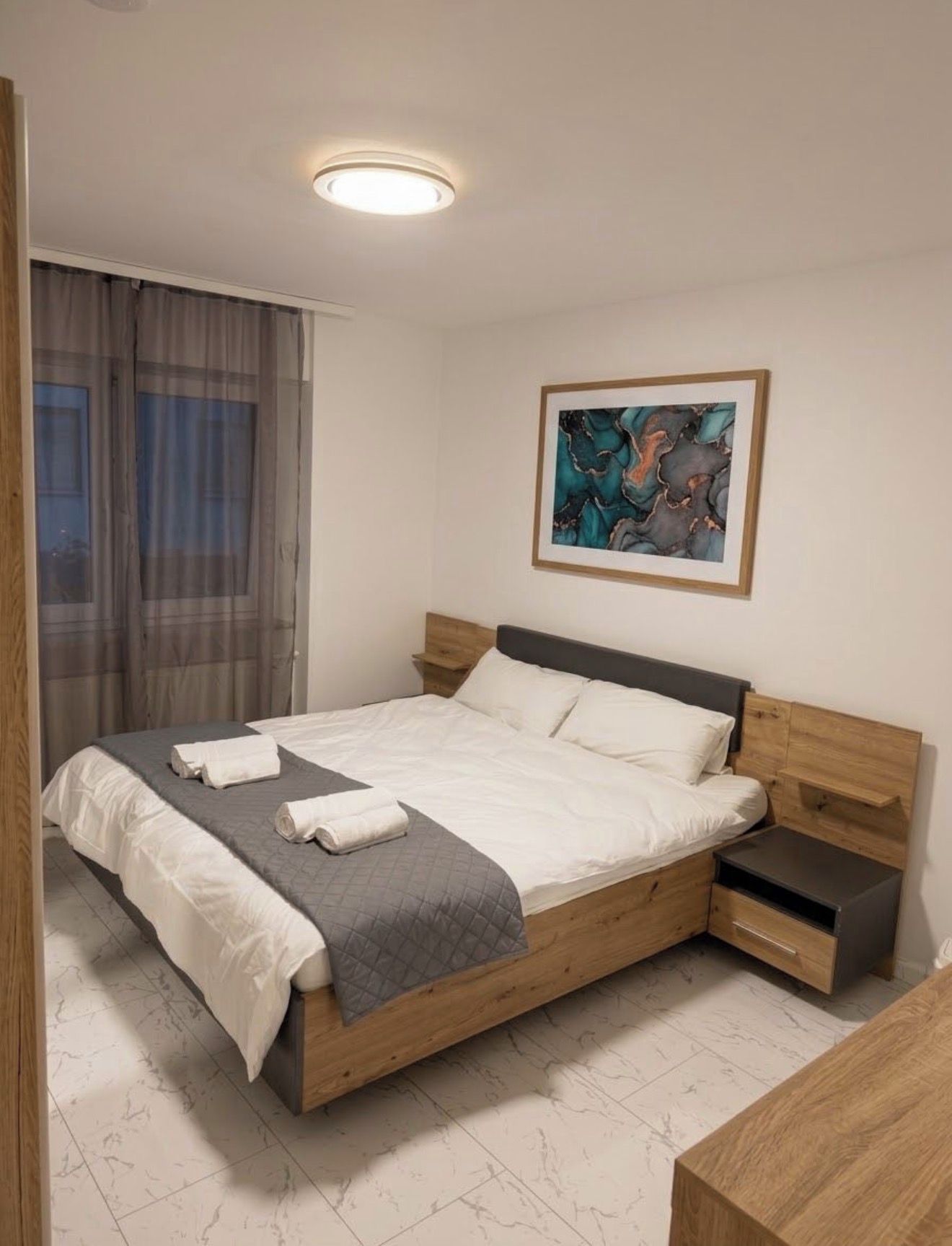 A modern bedroom with a bed, bedside tables, a window with curtains, and a framed abstract painting on the wall.