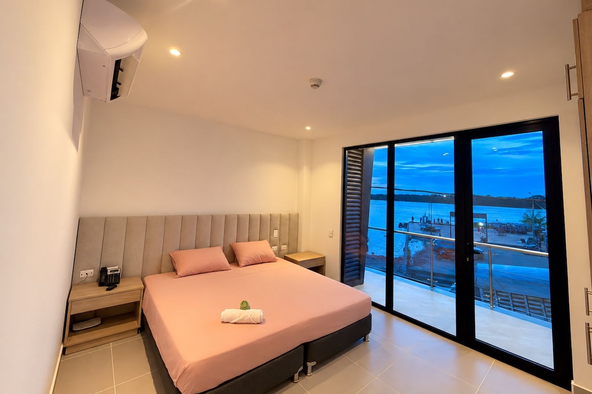 Un dormitorio moderno con cama, mesitas de noche, aire acondicionado integrado y un amplio ventanal con vista a un cuerpo de agua.