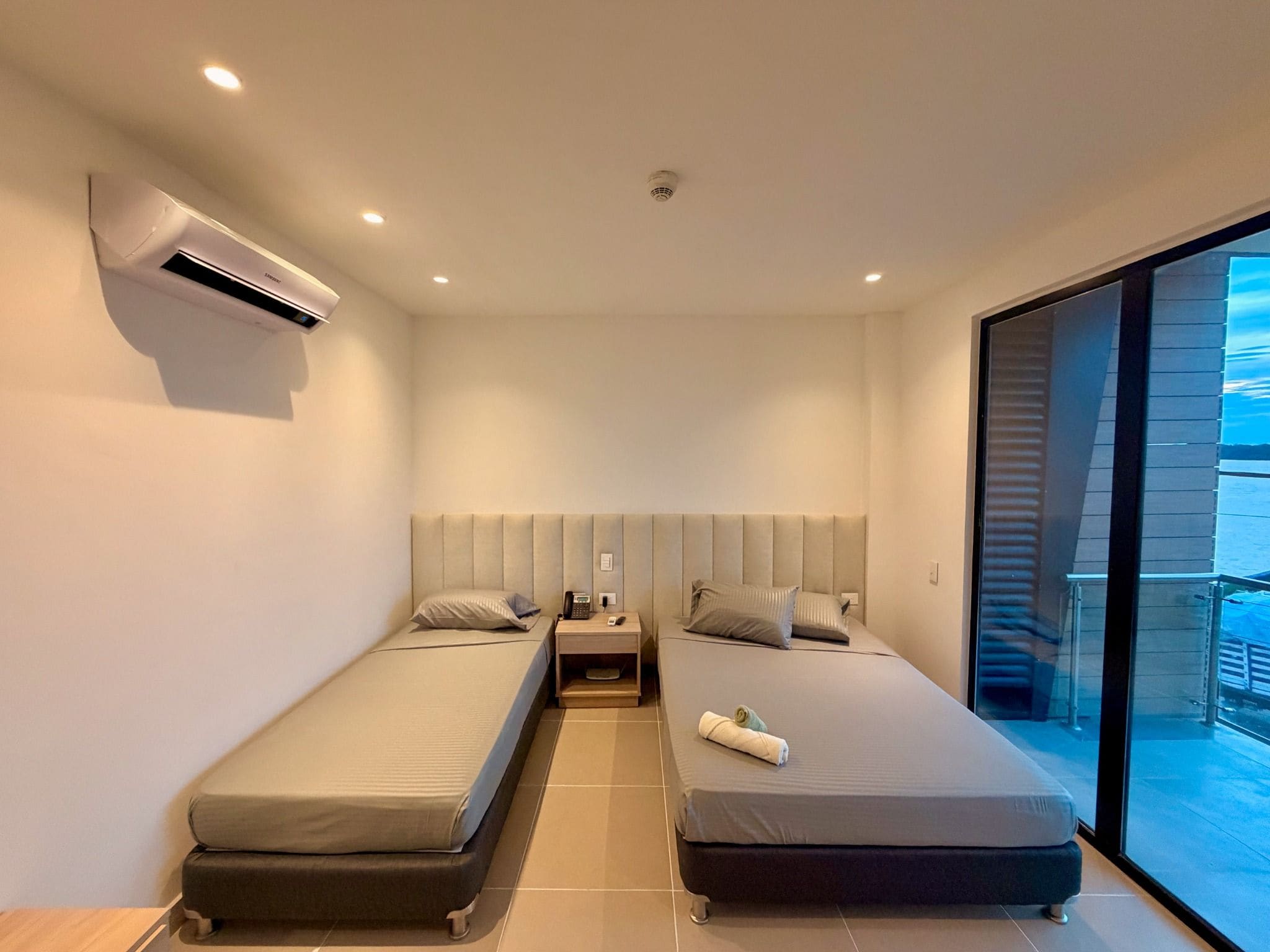 Habitación de hotel moderna con dos camas individuales, aire acondicionado integrado, mesita de noche con teléfono y lámpara, y un amplio ventanal con vistas.