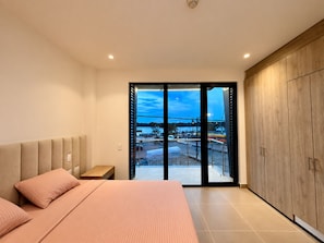 Un dormitorio moderno con cama, mesita de noche, armario de madera y vista a un paisaje urbano a través de una puerta de cristal.