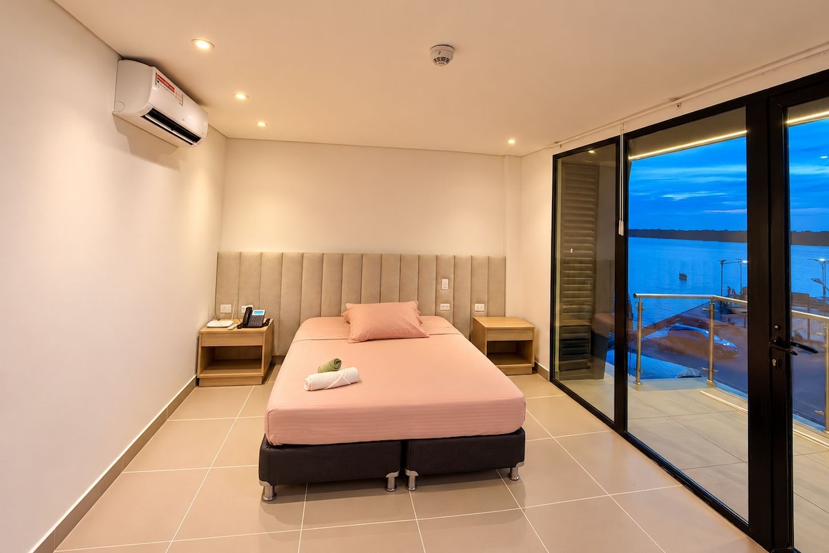 A modern bedroom with a large bed, a bedside table, a nightstand, an air conditioning unit, and a view of a balcony with a railing.