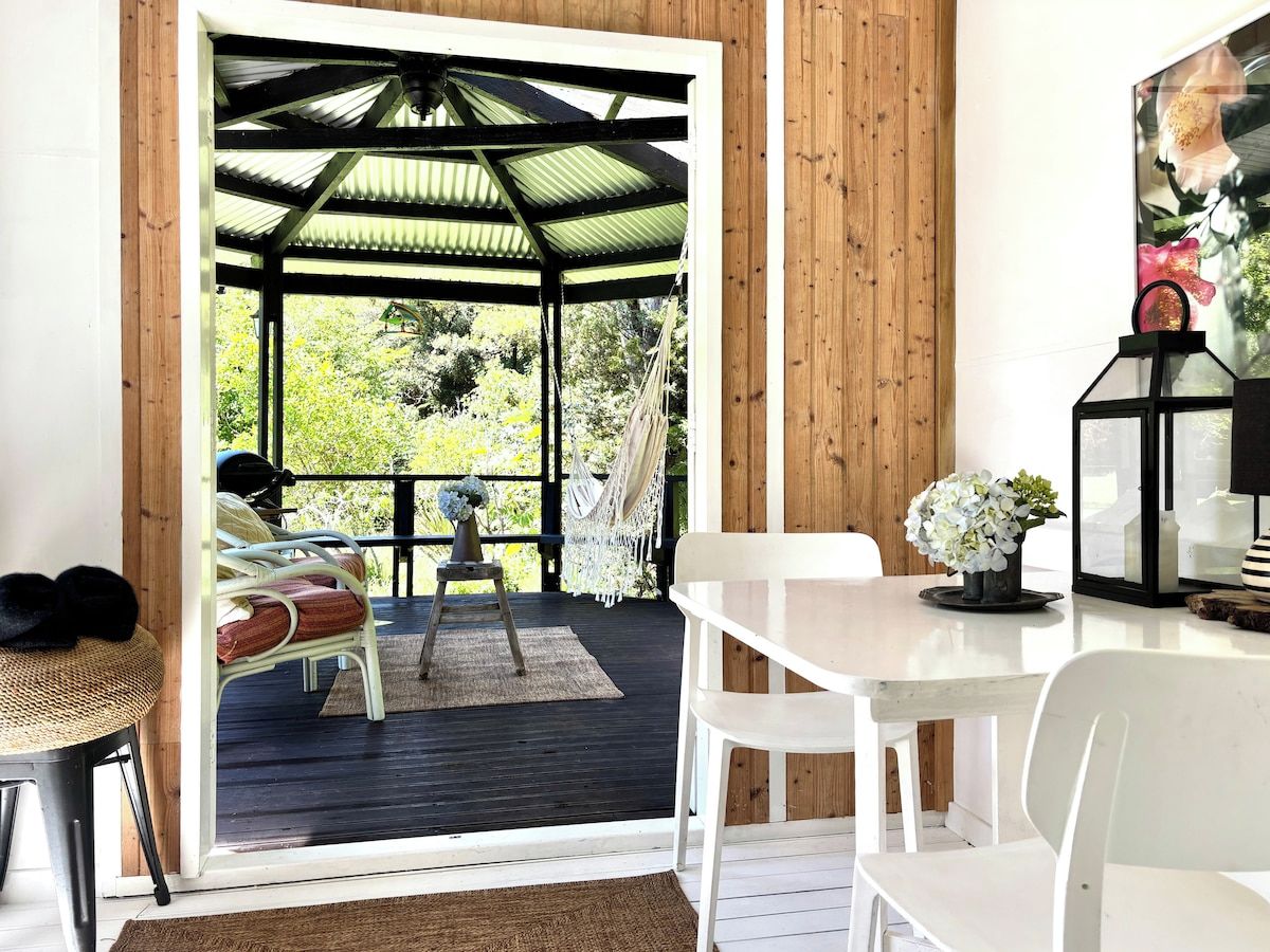 A room with a wooden floor, a white table, a white chair, a hanging chair, and a view of a garden through a glass door.