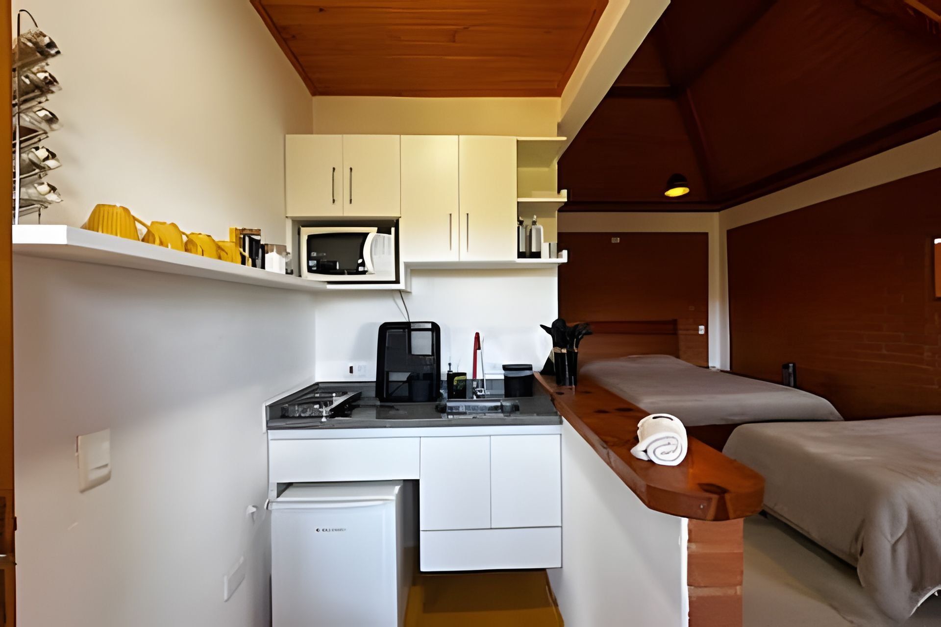 Une petite cuisine équipée d’un four à micro-ondes, d’une machine à café et d’un petit réfrigérateur. On y trouve également un banc en bois avec une serviette, ainsi qu’un lit avec une tête de lit.