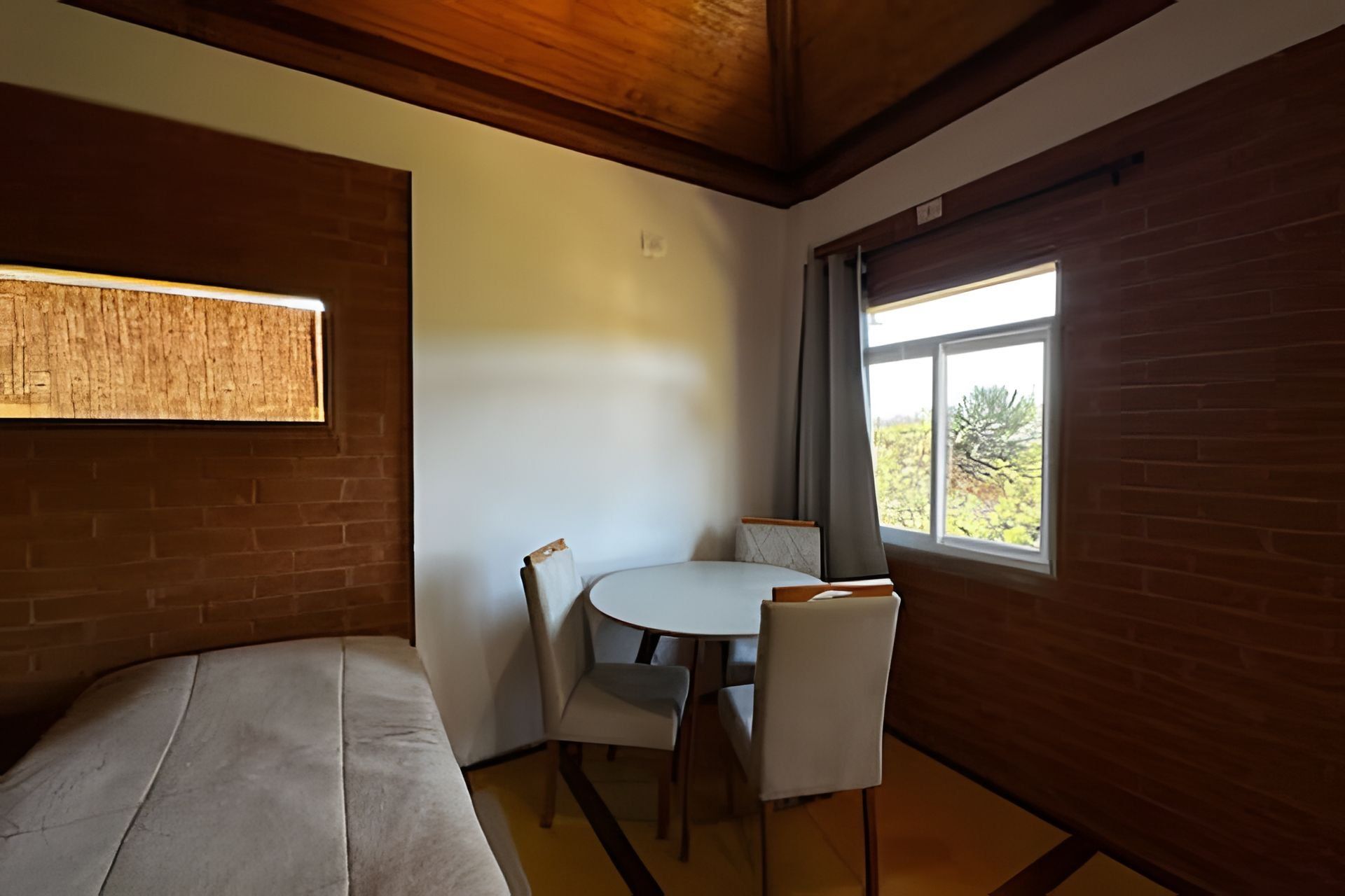 A room with a bed, a small table with chairs, a window with a view of greenery, and wooden beams on the ceiling.