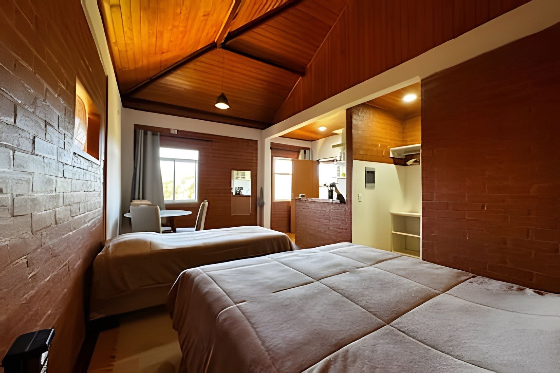 Quarto com duas camas, uma mesinha e uma cadeira. Há uma janela com cortinas, uma parede de tijolos e um teto de madeira.