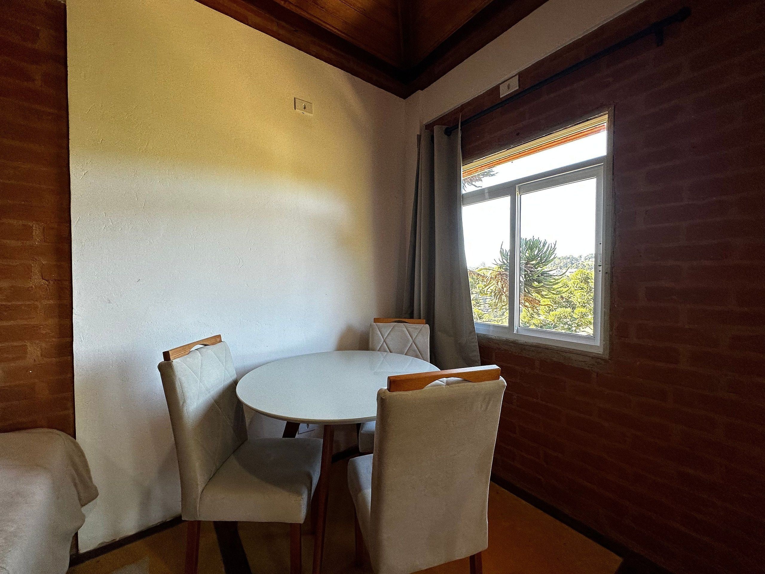 A dining area with a round table and four chairs, a window with blinds, and brick walls.