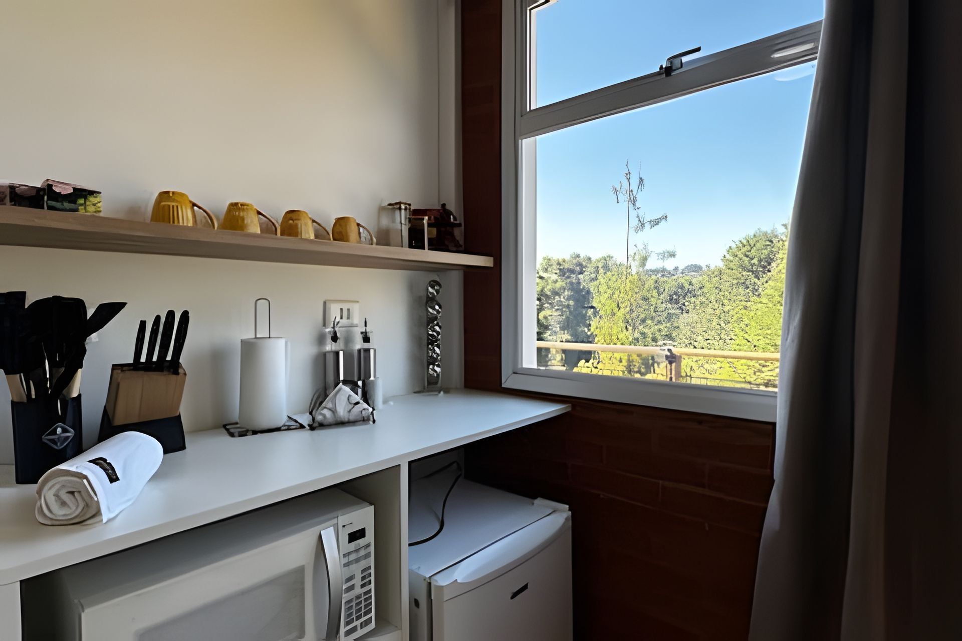 Una cocina con mostrador blanco, un refrigerador pequeño y una ventana con vista a árboles y una cerca.