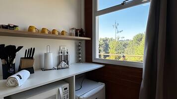 Una cocina con mostrador blanco, un refrigerador pequeño y una ventana con vista a árboles y una cerca.