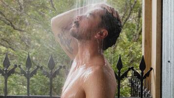 Eine Person steht unter einer Dusche, während Wasser auf sie fällt. Im Hintergrund sind ein Metallzaun und Grünpflanzen zu sehen.