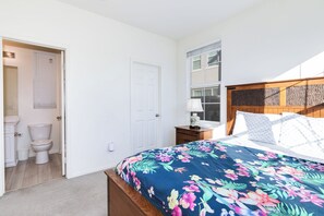Een slaapkamer met een bloemenprint sprei, een houten hoofdbord, een nachtkastje met een lamp en een raam met jaloezie ën.