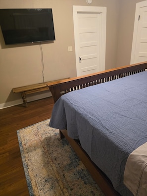 A bedroom with a bed, a bench, a television mounted on the wall, and two doors. - The Shaded Pecan Cottage (Terrell)