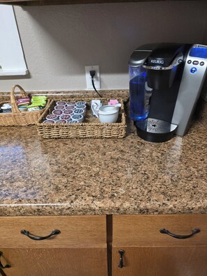 A kitchen counter with a coffee maker, a basket of coffee pods, a mug, and a wicker basket containing various items. - The Shaded Pecan Cottage (Terrell)