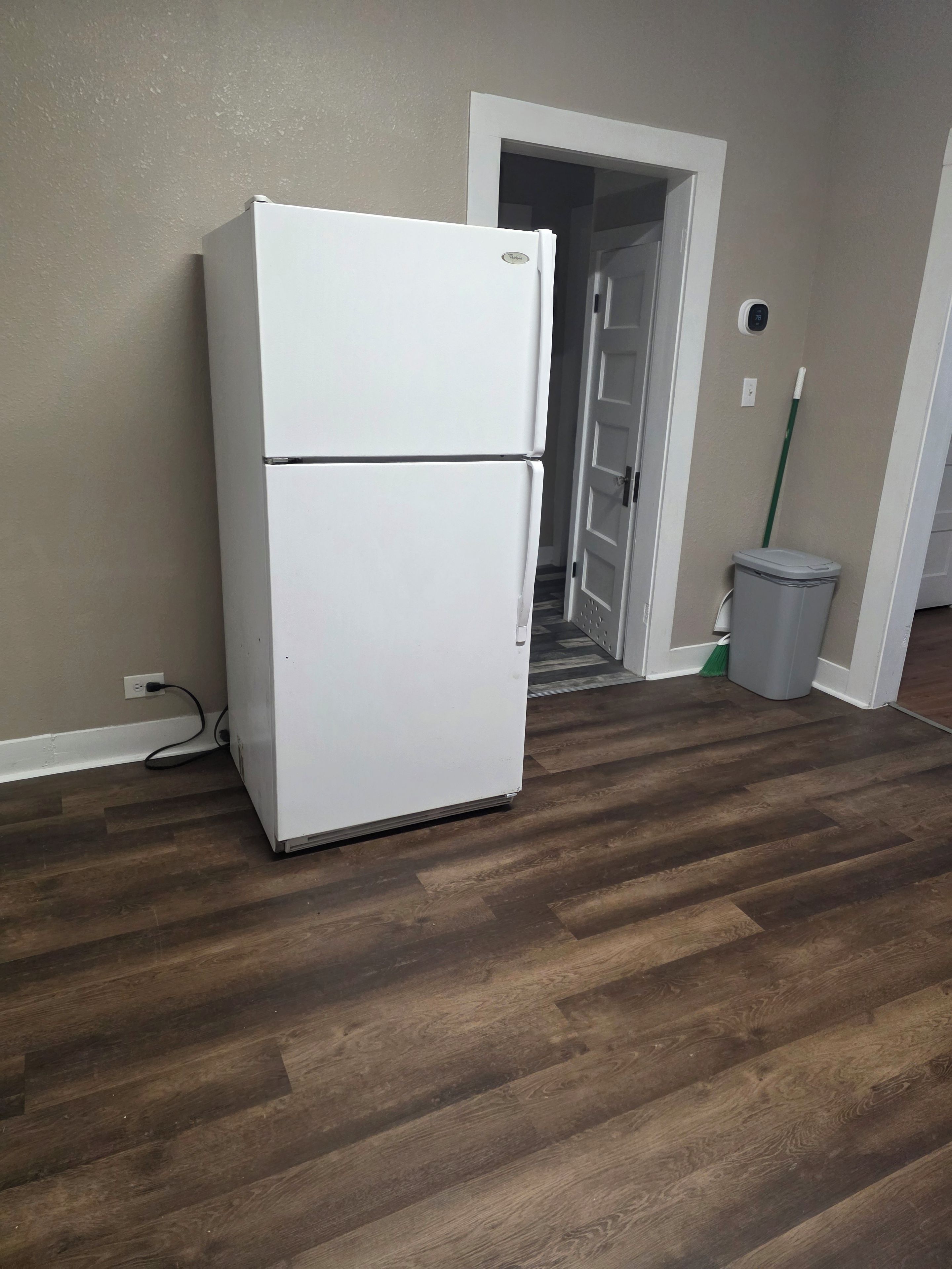 Un réfrigérateur blanc se trouve dans une pièce dont le sol est recouvert de parquet et qui possède une porte donnant sur un couloir.