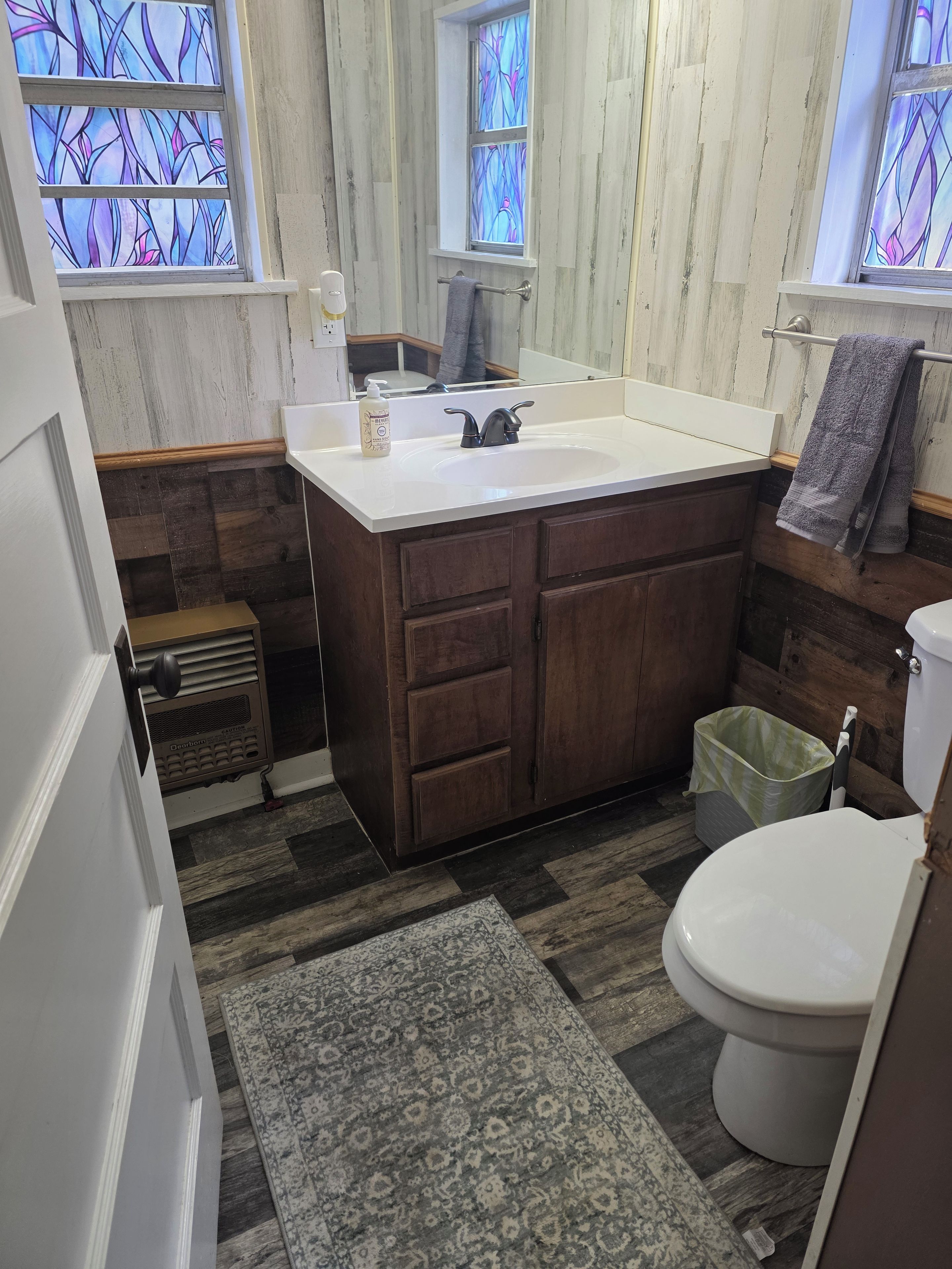 A bathroom with a wooden vanity, a white sink, a toilet, and a green trash bin.