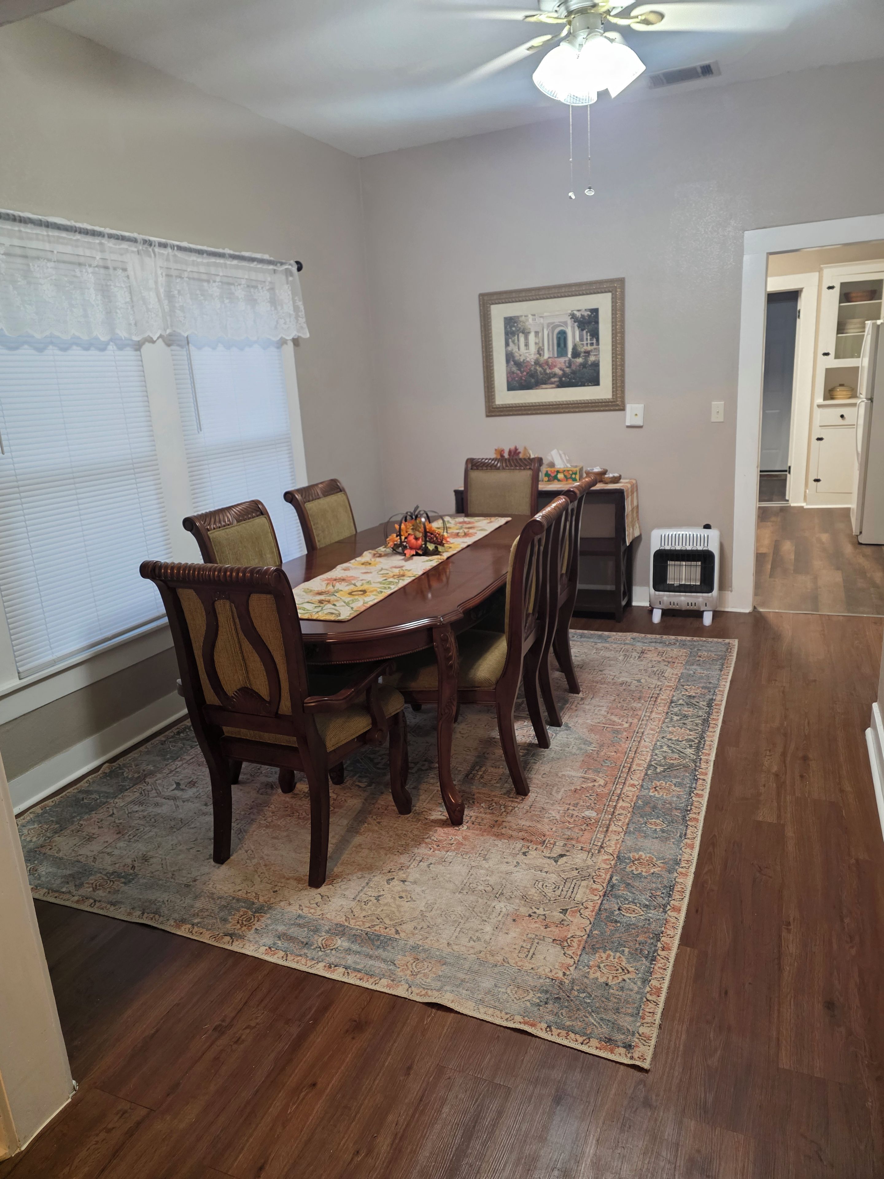 A dining area with a wooden table, six chairs, a patterned rug, a ceiling fan, and a framed picture on the wall.