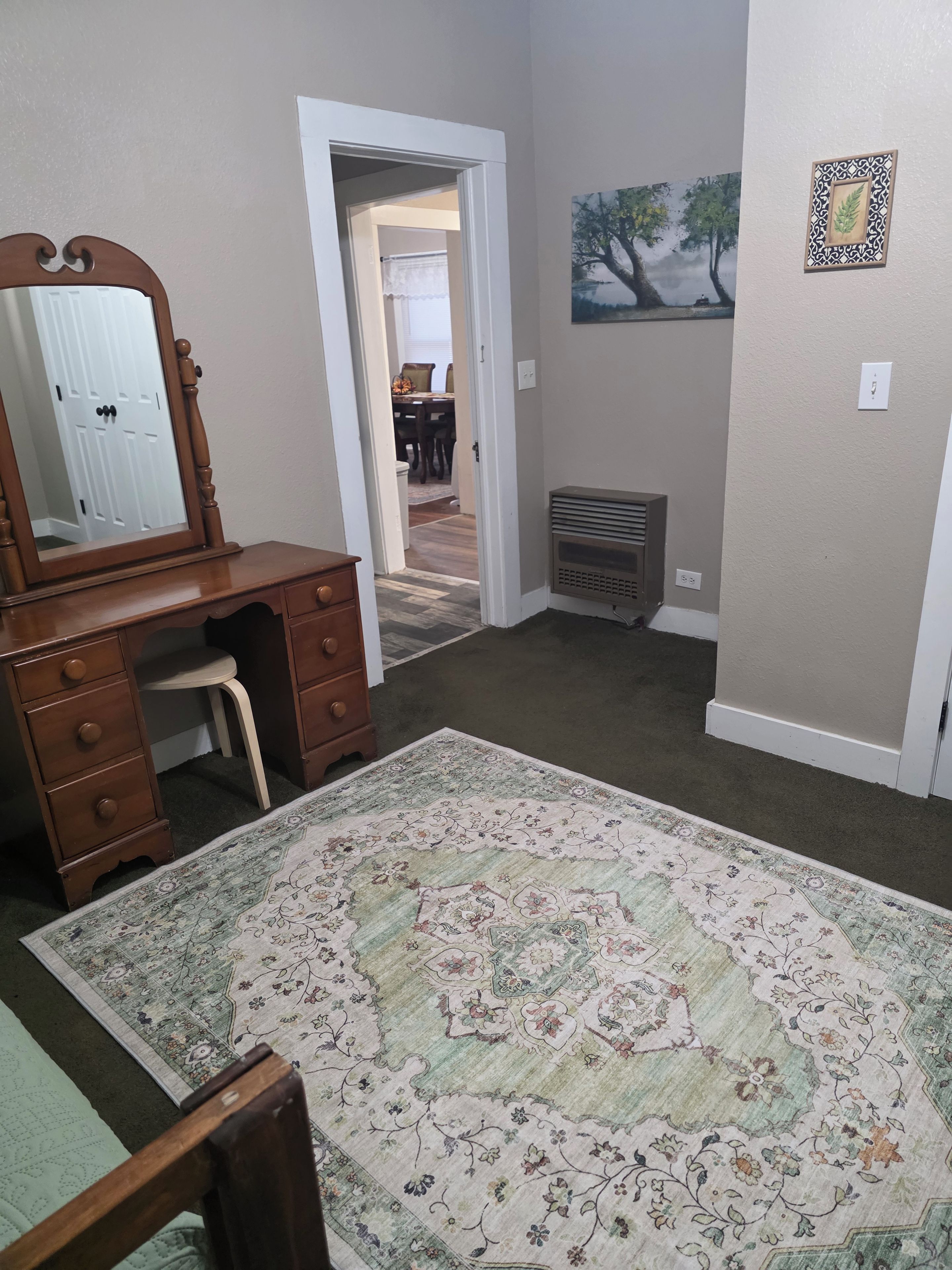 A room with a wooden dresser, a large mirror, a patterned rug, a radiator, and a painting on the wall.