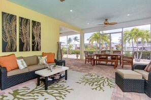 A spacious living area with a sofa, coffee table, and a dining set. There are large windows providing a view of palm trees and a building. - Sunny Daze~Private Pool Home, 45ft of Dockage, minutes from the beach! (Marathon)