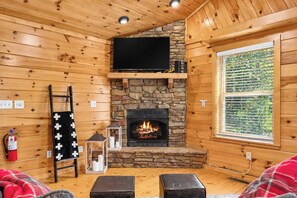 Ein gemütliches Blockhaus-Interieur mit Kamin aus Stein, Holzwänden, einem darüber montierten Fernseher und einem Fenster mit Jalousien.