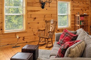 A cozy cabin interior with a rocking chair, a gray sofa with plaid cushions, and a deer head mounted on the wall. - Stonebridge Retreat | Hot Tub • Near Everything! (Sevierville)