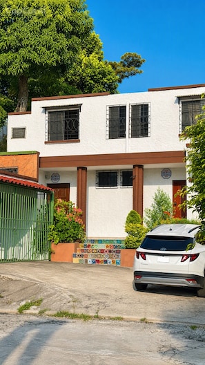 A two-story building with a white facade, multiple windows with metal bars, and a green gate. - La Casa del Pasaje (San Salvador)