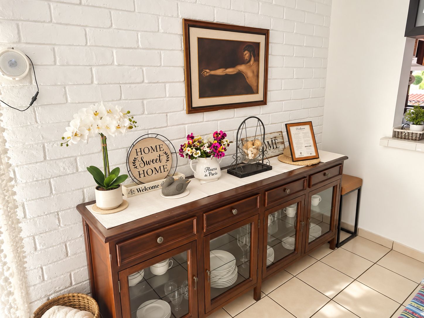 Une cuisine avec un mur en briques blanches, un tableau encadré, une pancarte de bienvenue, une orchidée en pot et un meuble avec des portes en verre.