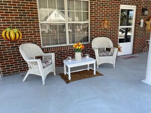 A brick building with a porch featuring two wicker chairs, a small table, and a vase with flowers.