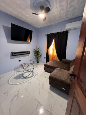Ein Wohnzimmer mit einem Flachbildfernseher, einem Deckenventilator, einem Glastisch, einer Couch und einer Topfpflanze.