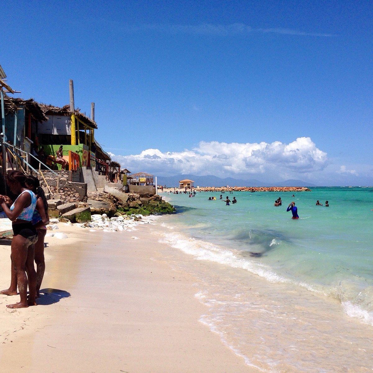A beach with clear water, people swimming, and buildings in the background.