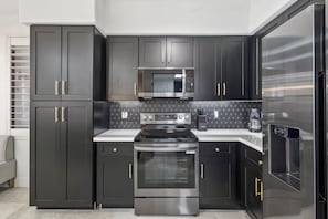 A modern kitchen with dark cabinetry, a stainless steel refrigerator, an oven, a microwave, and a coffee maker. - Black & Gold Luxe | No Stairs | Pool & Spa (Scottsdale)