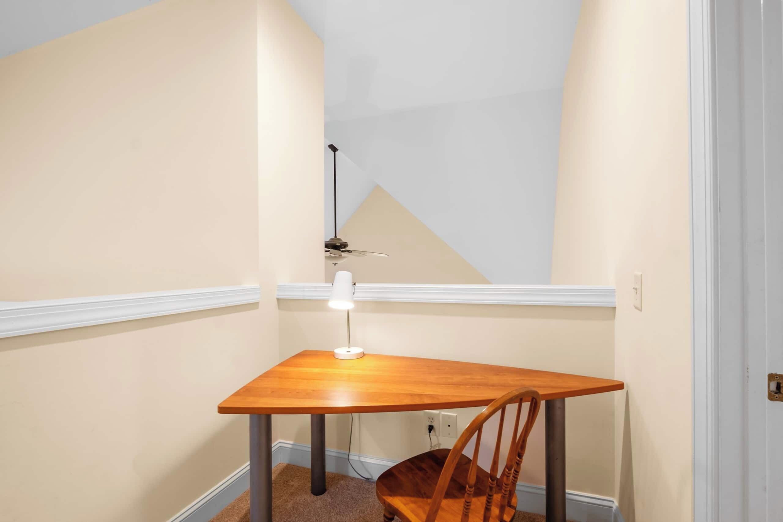Un rincón pequeño y bien iluminado con una mesa de madera y una silla, un ventilador de techo y una pared blanca con zócalo.