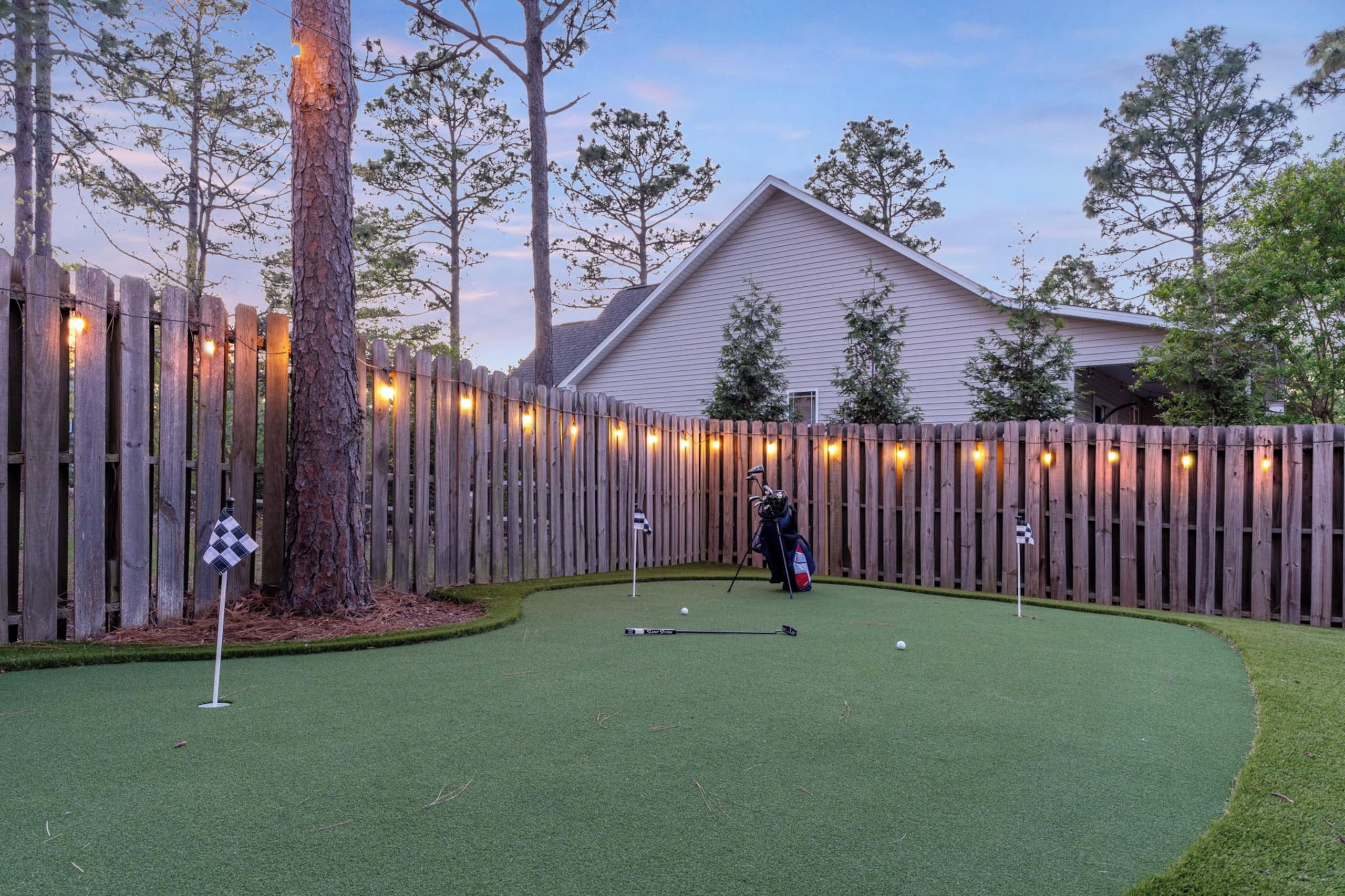 Un campo de mini golf con una cerca de madera, una casa al fondo y un bolso de golf en el área de salida.